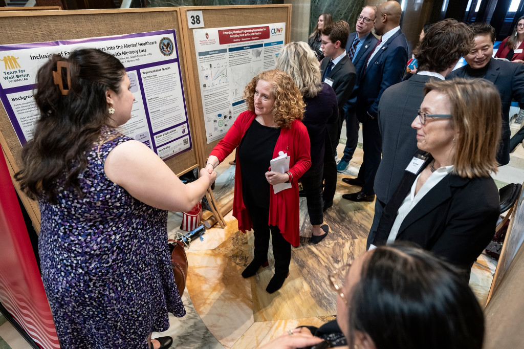 Two women shake hands in front of a poster board, another person looks on.