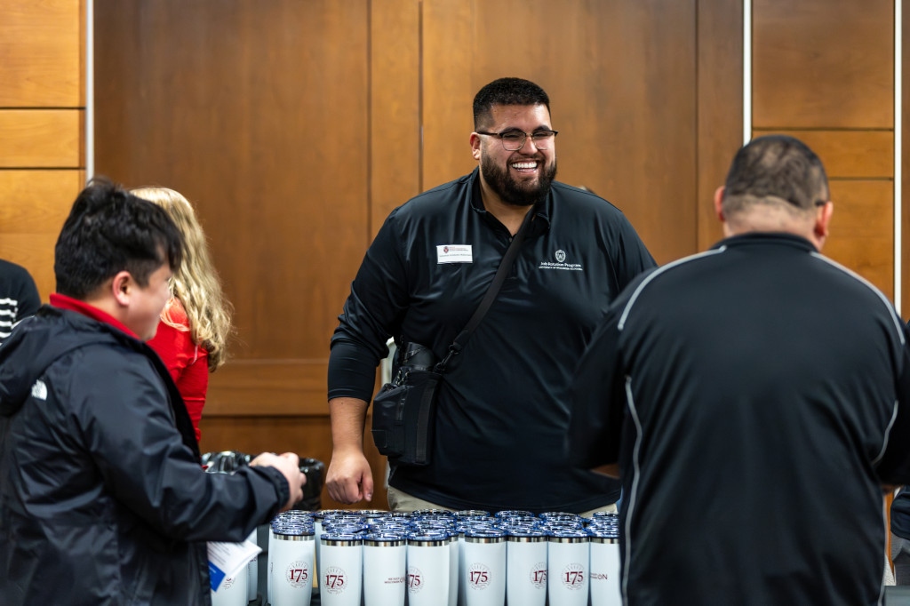 A person standing behind a table smiles and talks with other people.