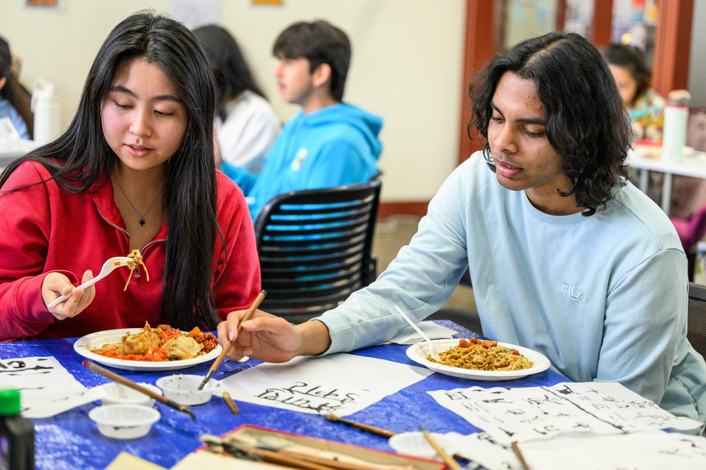 People sit at a table and do calligraphy, with plates of food nearby.