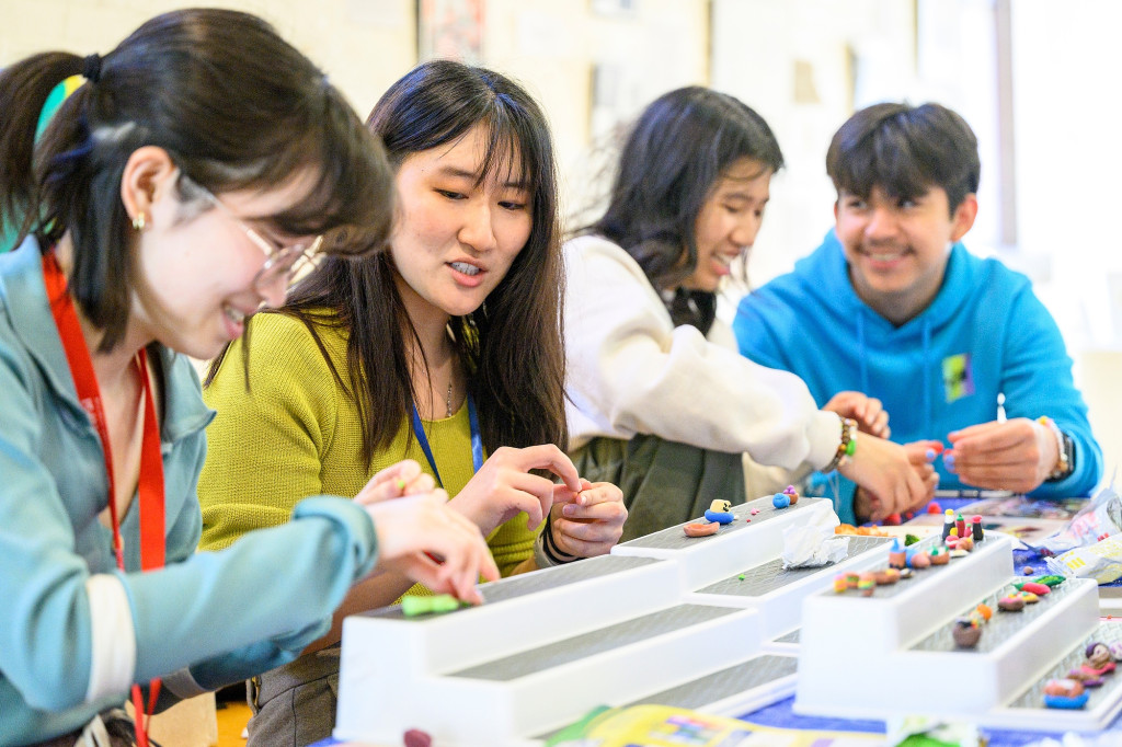 Four people are lined up at a table, making miniature art and talking.