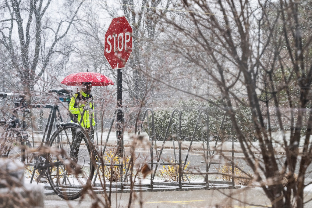 A person with a yellow raincoat and red umbrella walks on a sidewalk with snow falling around them.