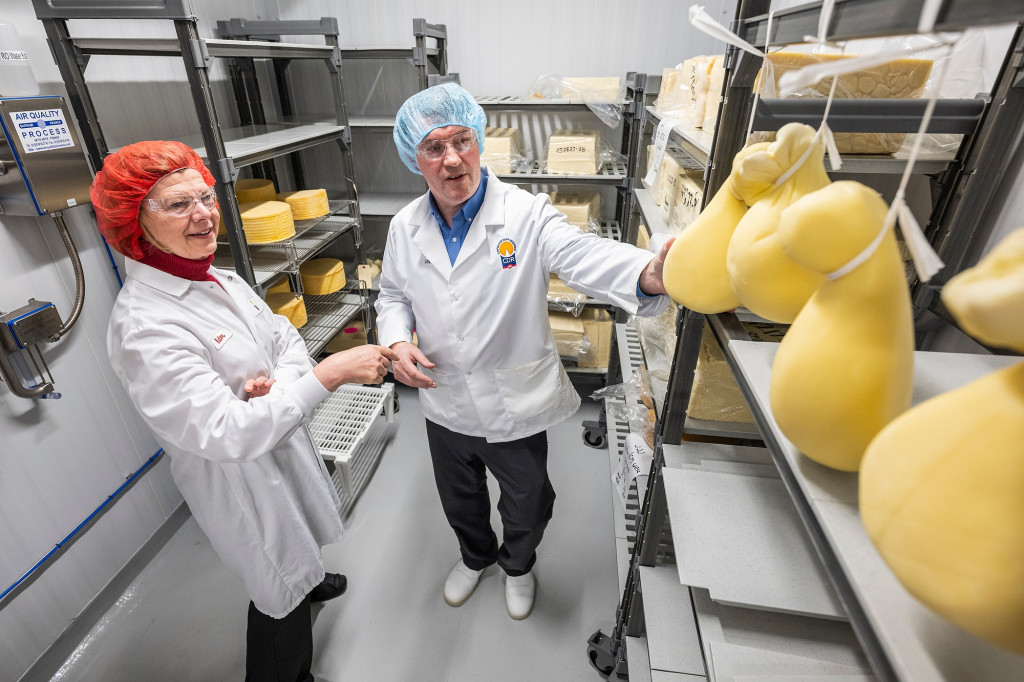 Two people talk next to shelves storing lumps of cheese.