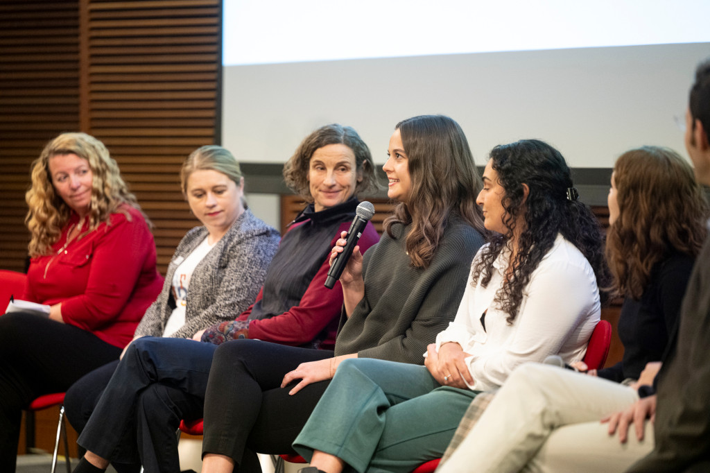 A row of panelists sit on a stage; one speaks into a microphone.