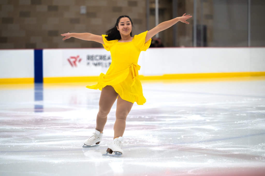 A woman in a yellow dress skates on an ice rink.