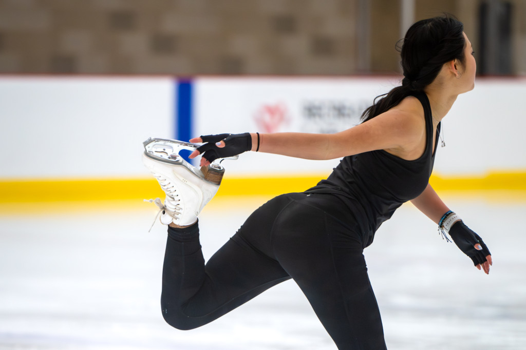 A woman reaches back and clasps her skate blade while skating on the other leg.