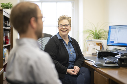 Clare Hun smiles as she sits at her desk while speaking to a colleague