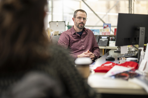 Teddy Kaul sits at a computer desk while listening to colleagues.