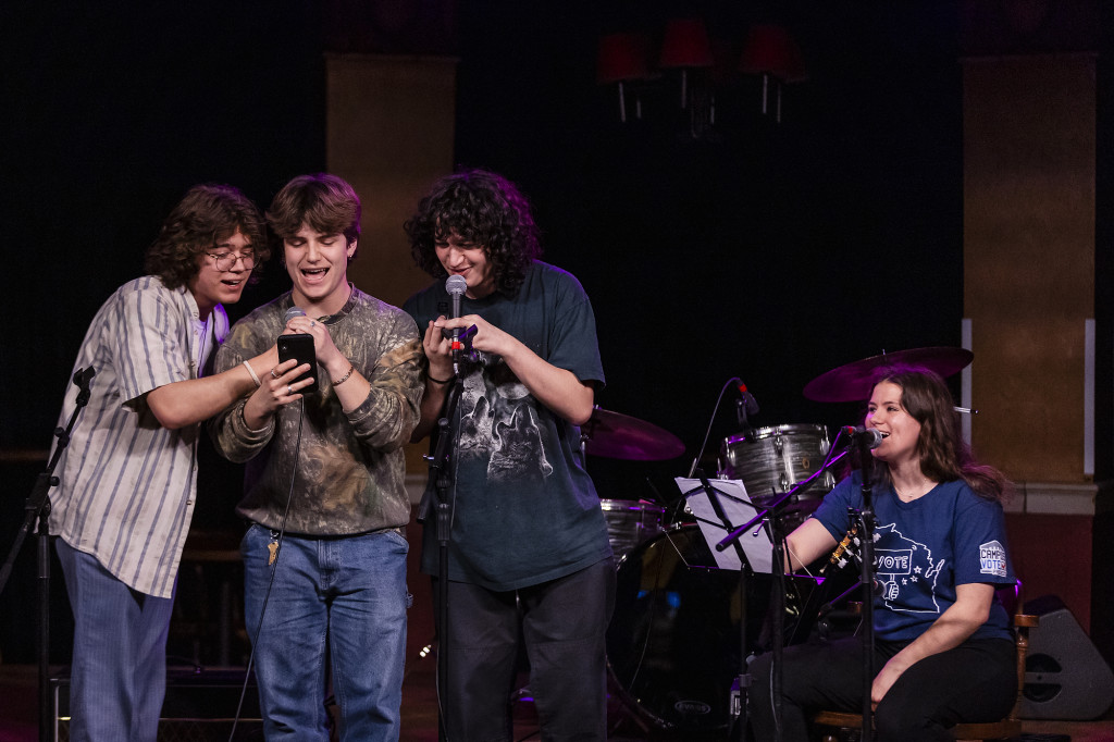 A photo of several singers on stage, one looking at a cell phone.