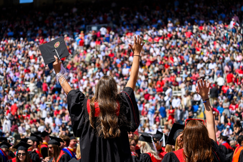 Graduates in academic robes stand and wave their hands.