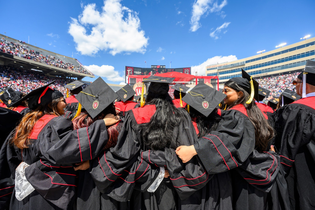 People in black commencement robes stand, arms around each other, and sing.