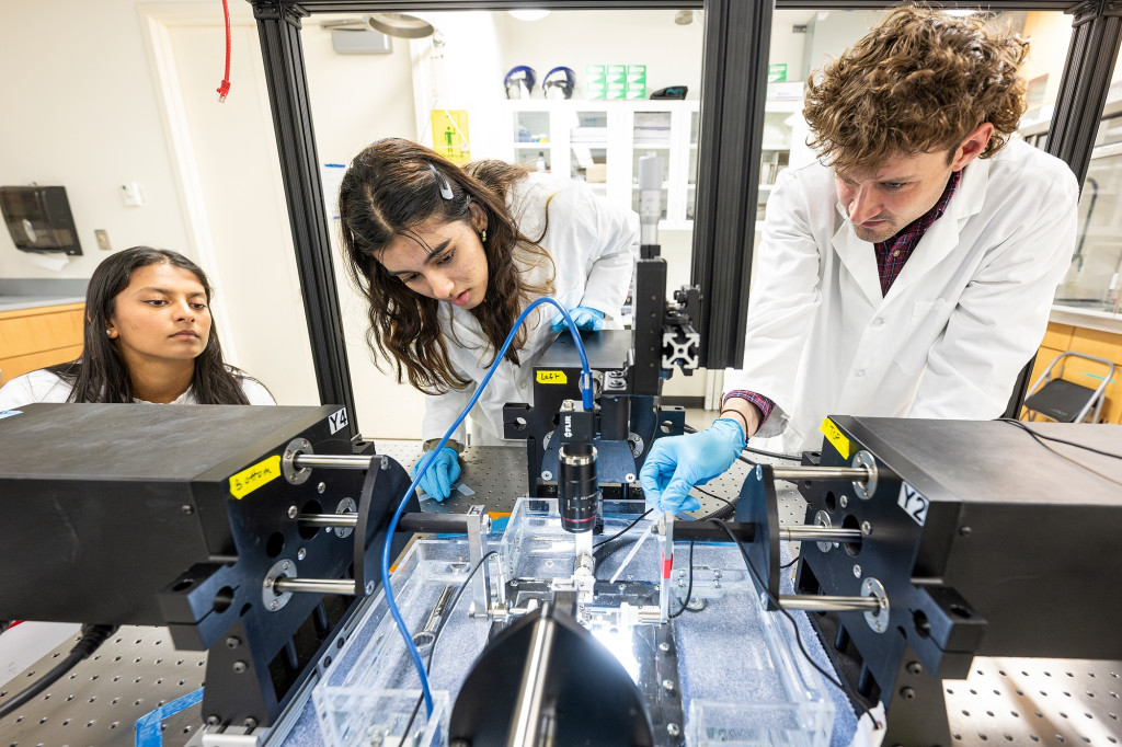 Three people wearing white lab coats bend over lab equipment as they work on an issue.