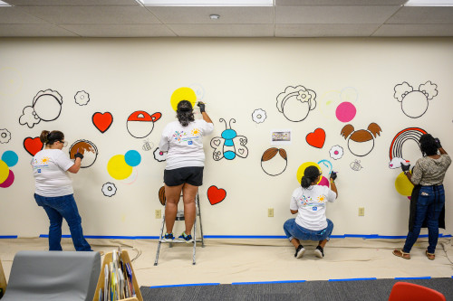 Four people face away from the camera as they paint a colorful mural on a white wall.