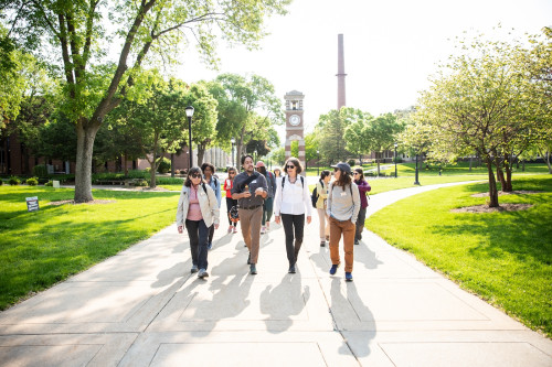 People walk on a concrete pathway across a campus with buildings and grassy fields.