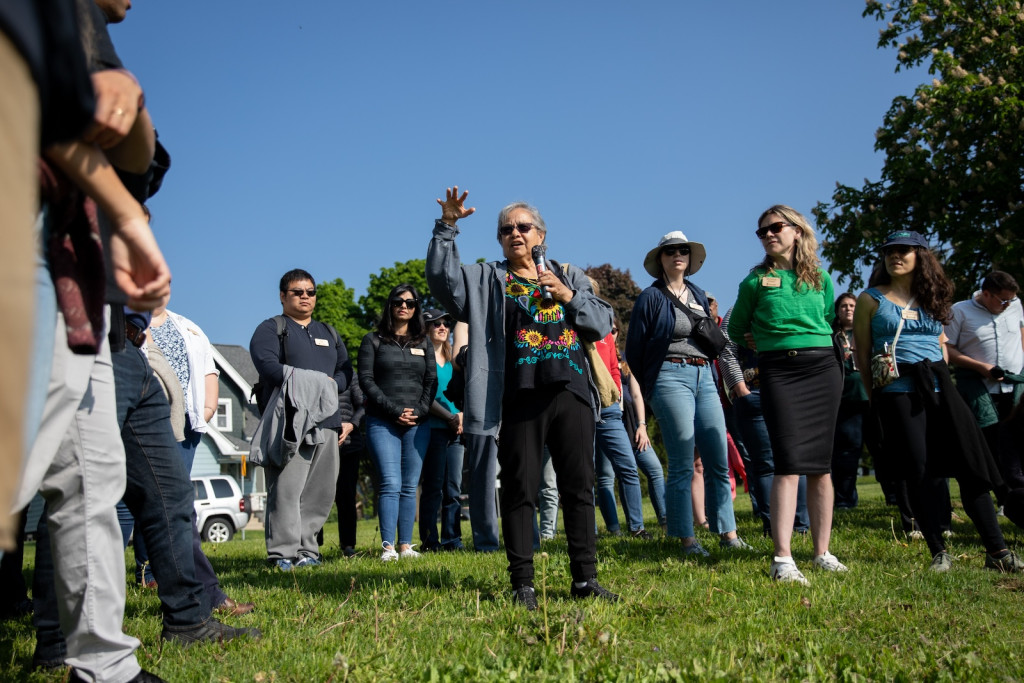 A man speaks to a group of people, all of them standing in a park.