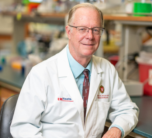 Photo of Allan Brasier smiling to the camera. He is wearing a white lab coat with a UW–Madison crest. His arm is leaning on a countertop in a laboratory.