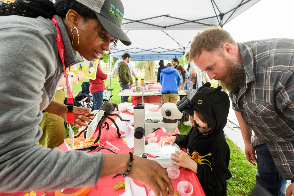 A man and young girl step up to a display table where a woman is giving a demonstration examining insects under a microscope.