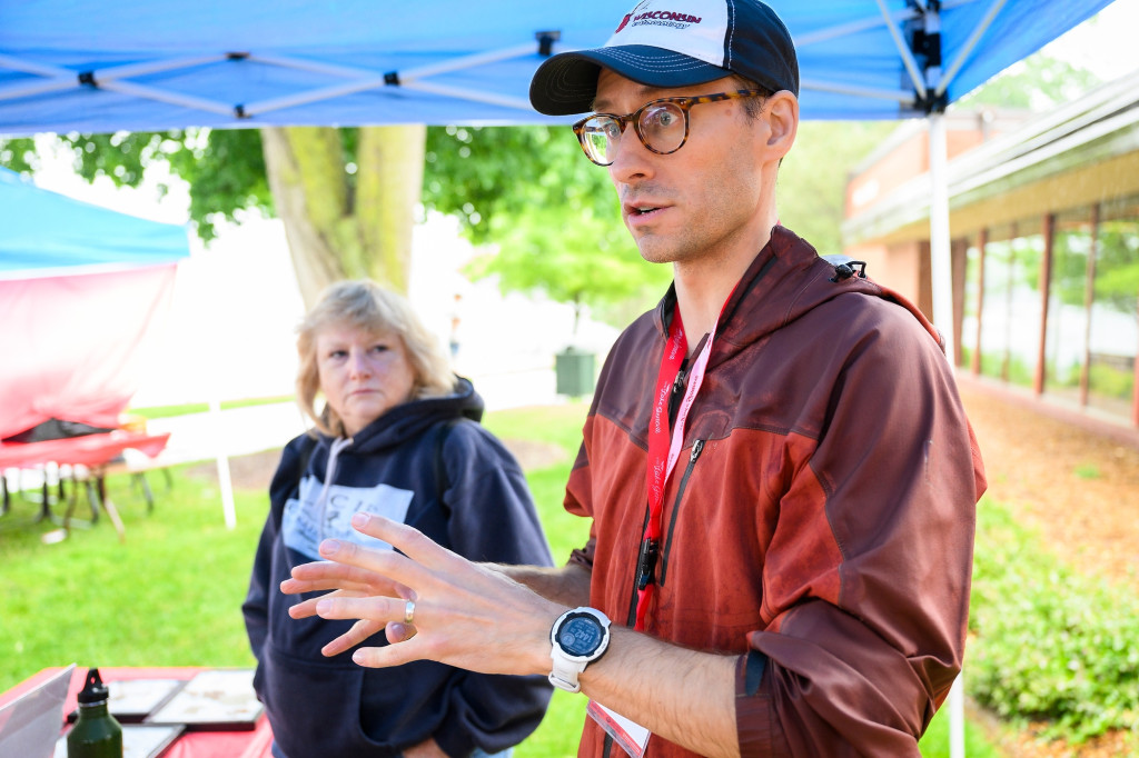 A man standing outdoors under a popup tent speaks and gestures with his hands. A woman behind him listens in.