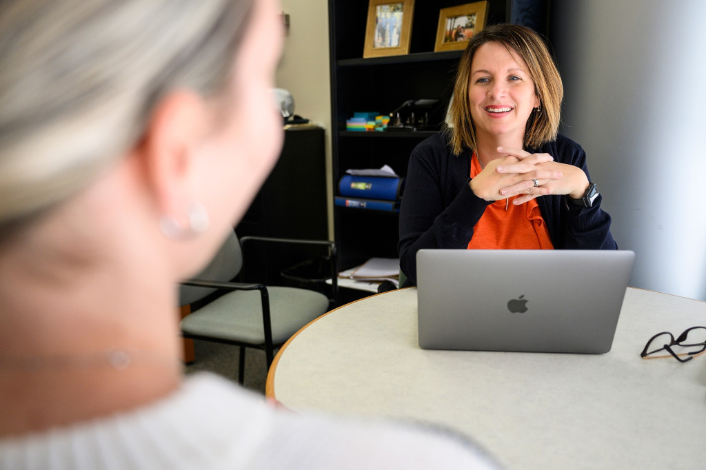 A woman facing the camera smiles and talks with a person in the foreground,