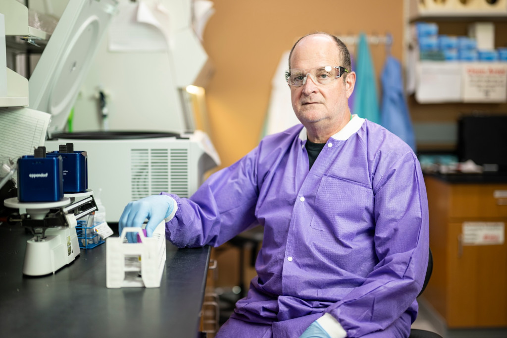 A man in a purple lab coat works with some test tubes.