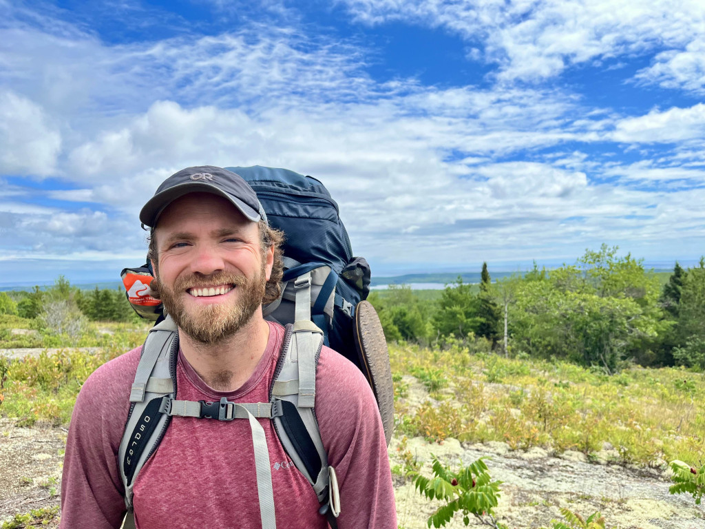 A man standing in a forest setting smiles at the camera. He wears a backpack and hiking clothes.