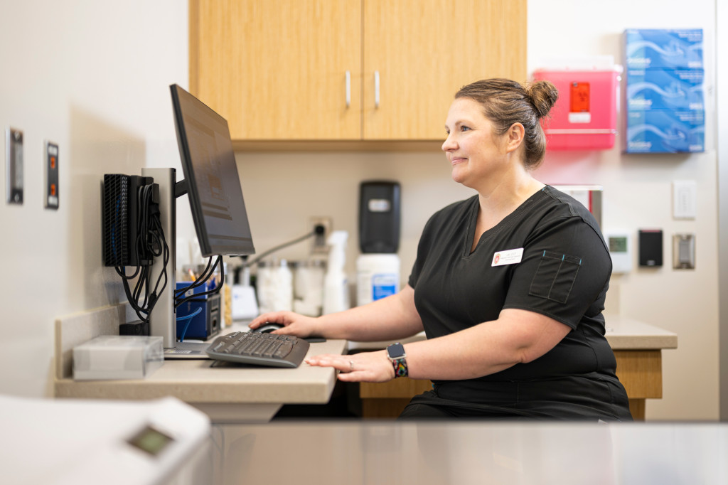 A woman in lab clothes is shown sitting at a desk, working at a computer.