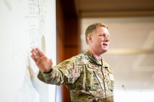 Zach Morris, wearing U.S. military fatigues, stands to give a presentation.