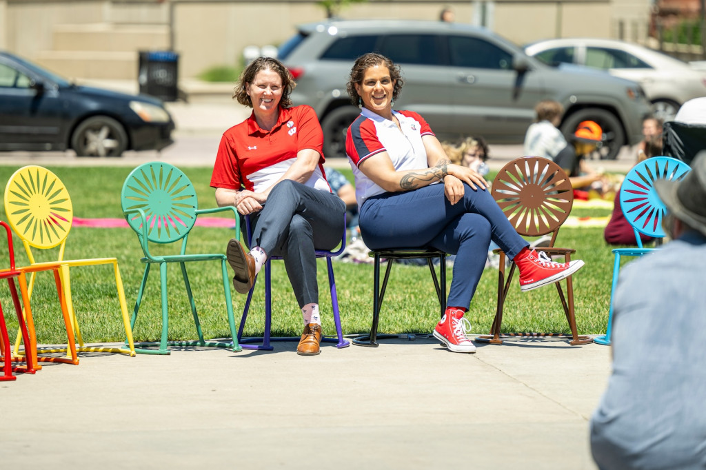 Two women sit in two chairs in a row of colorfully painted chairs.