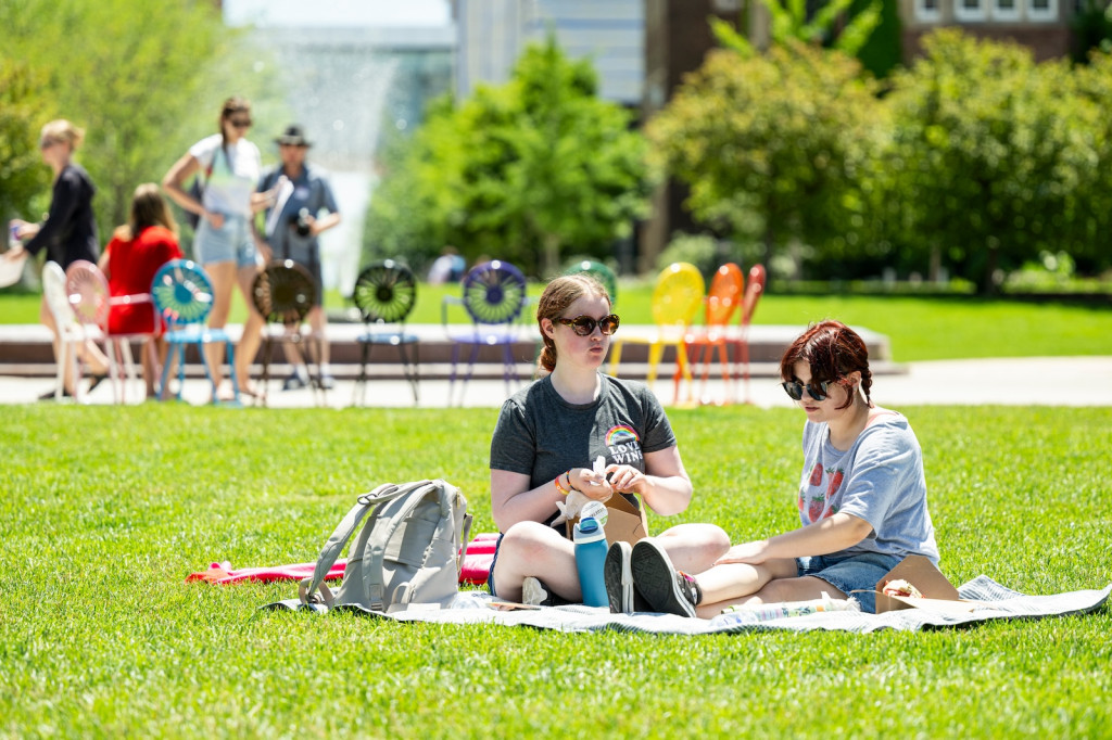 Two people sit on a blanket eating a picnic lunch.