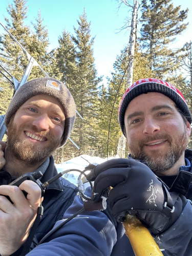 Two men in winter coats and hats pose in a forest covered in snow.