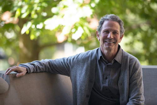 Person sitting on a concrete bench outdoors, smiling at the camera. They are wearing a gray cardigan over a checkered shirt. Trees with green foliage blur softly in the background. 