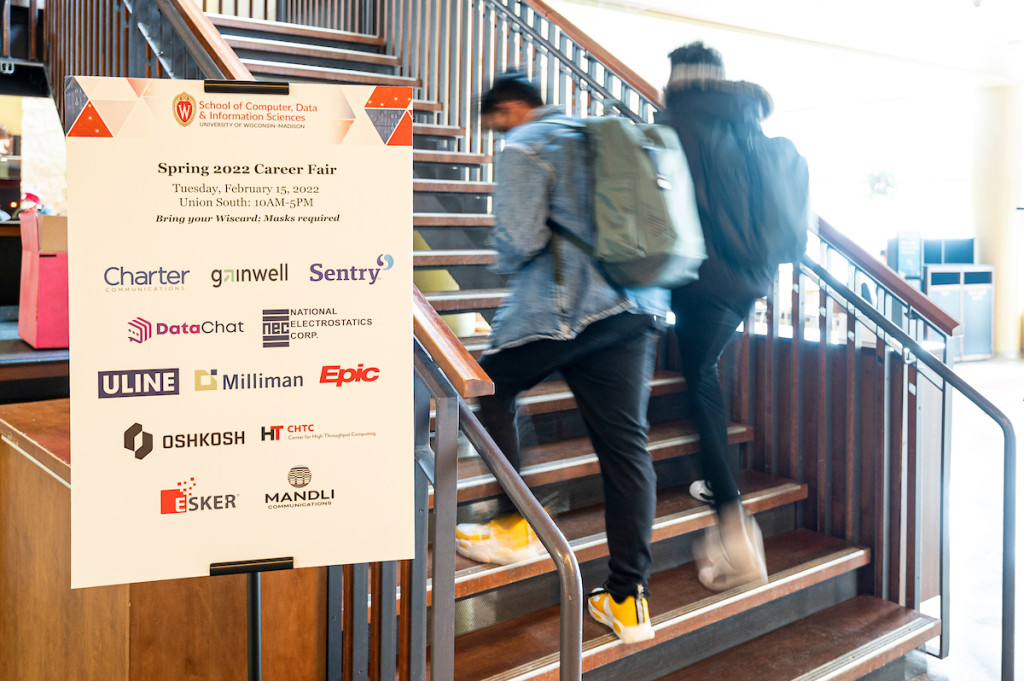 Two students walk up stairs as a sign listing employees is in the foreground.