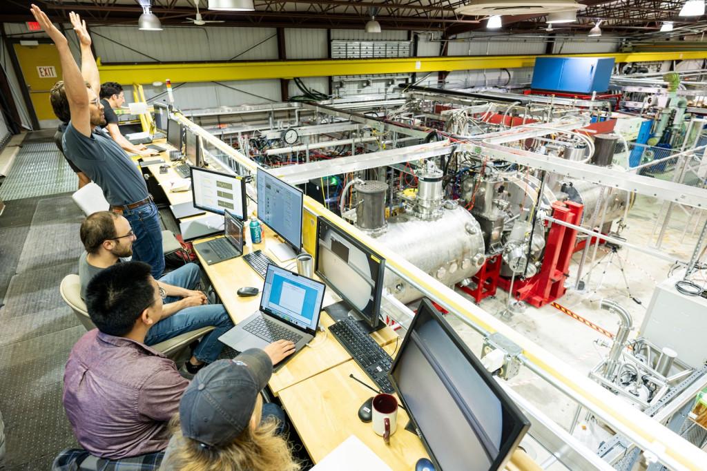 A number of people sit at a control panel overlooking equipment, one of them stands and raises his hands in celebration.