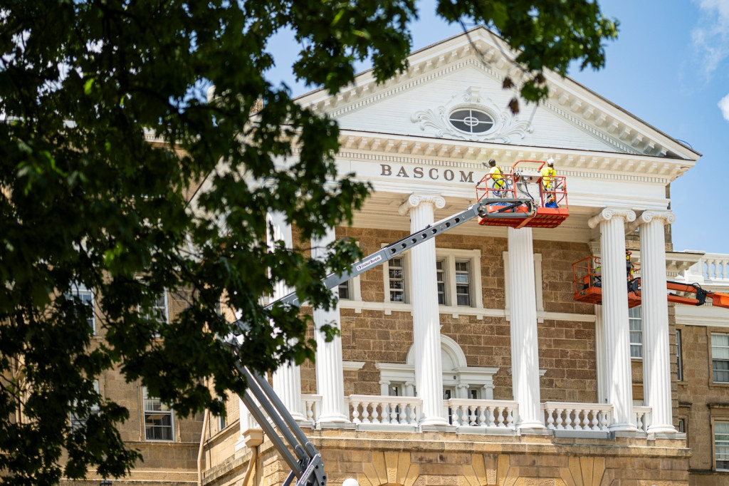 Workers clad in yellow safety vests paint the nameplate of Bascom Hall as they stand in lifts.