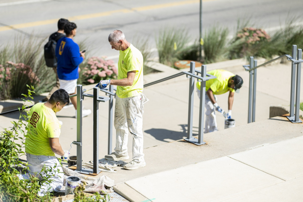 Workers in yellow safety shirts paint metal railings plante din concrete.