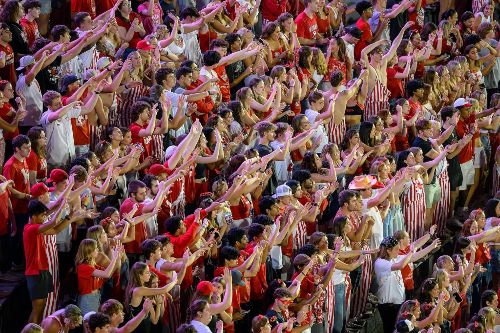 People in a stadium stands wearing red and white Badger gear wave their hands.