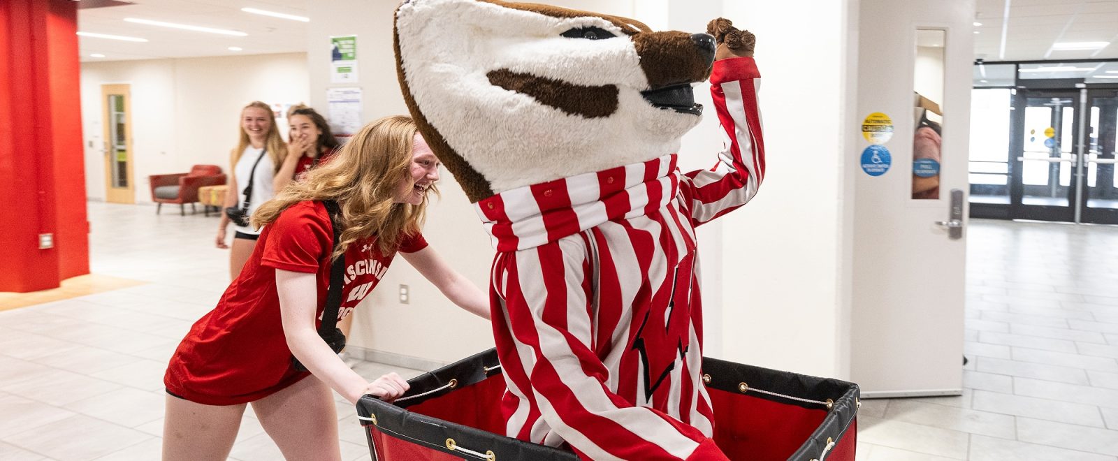 UW-Madison mascot Bucky Badger rides in a big red cart inside of a residence hall. He is being pushed by a student wearing a red Wisconsin Badgers t-shirt.