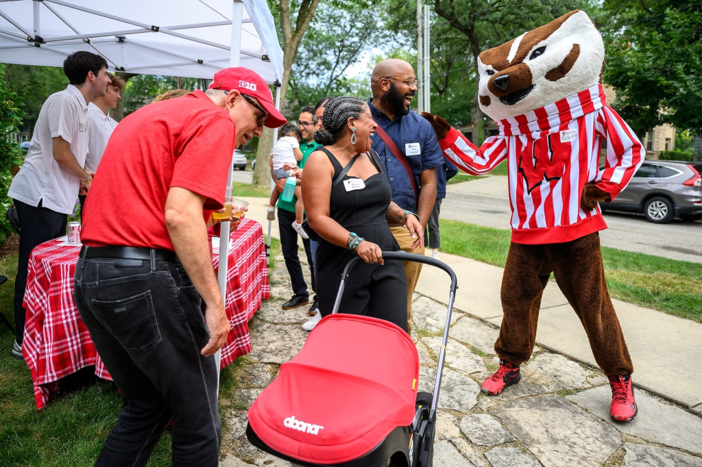 A woman pushing a baby carriage talks to Bucky Badger as another person admires the baby.