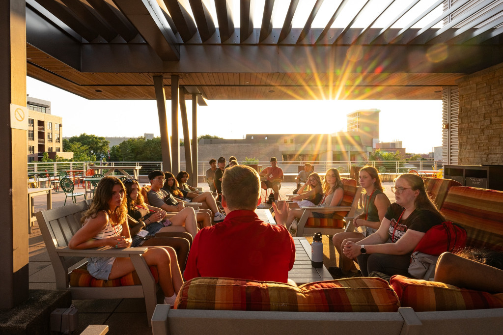 A group of college students sit on patio furniture talking as the sun goes down.