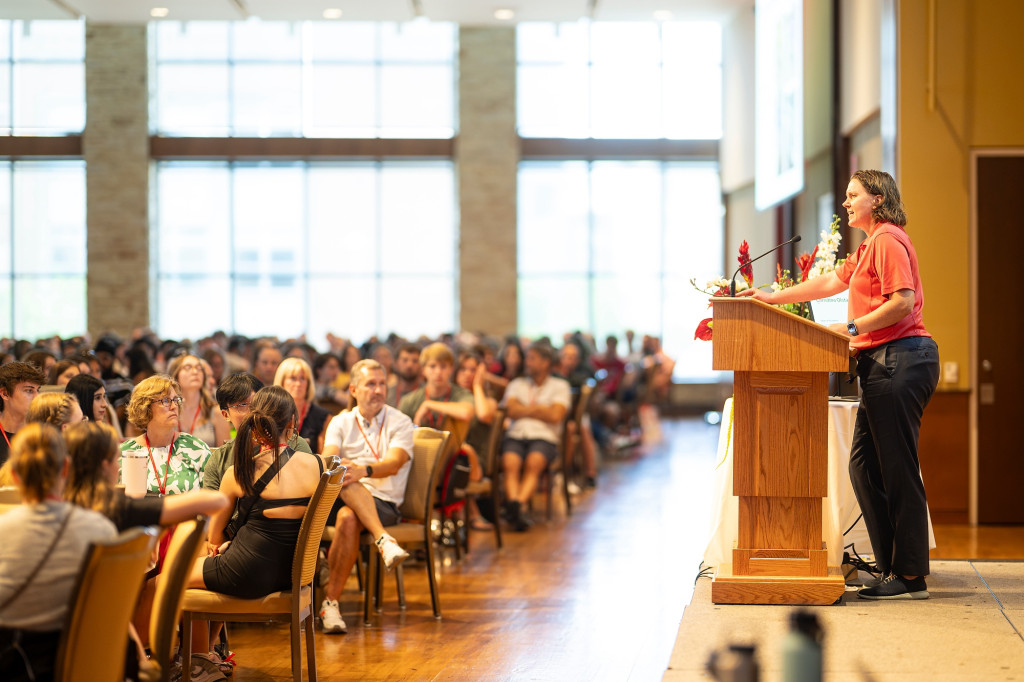 Side view of a person standing at a podium in a sun drenched room addressing a large seating group or parents and college students.