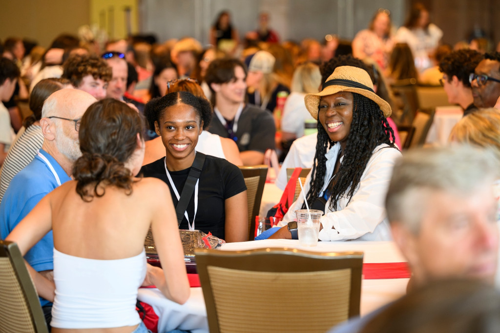 A group of students sit together in a crowded dining hall. They smile as they talk with one another.