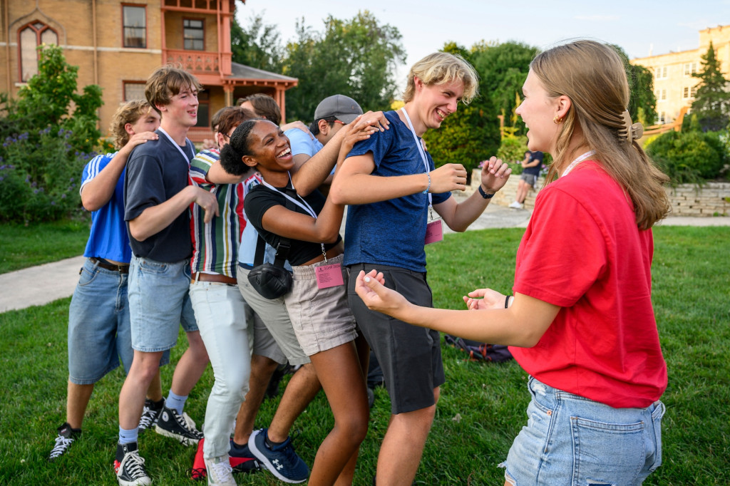 A group of college students laugh while standing in a line with their arms on the shoulders of the person in front of them.