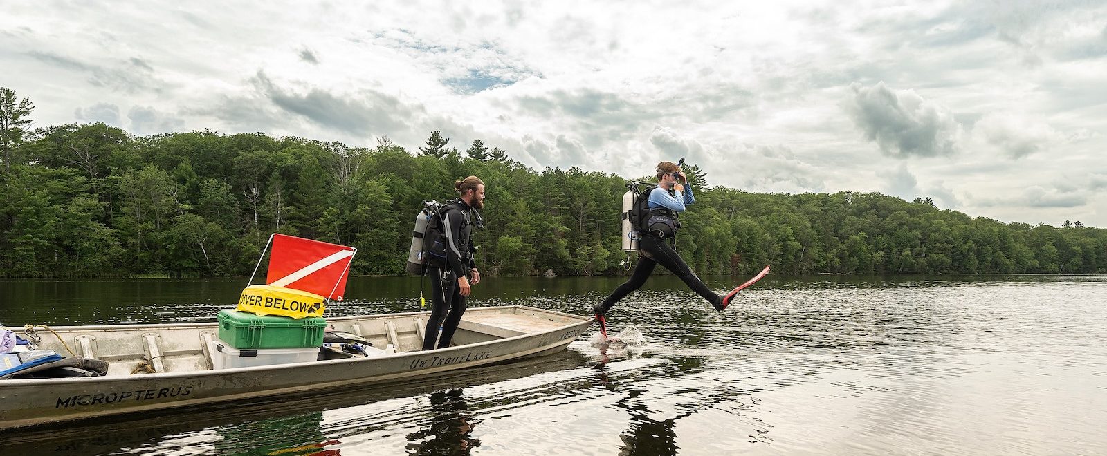 A person in scuba gear steps off the side of a boat into a Northwoods lake. A second person in the boat gets ready to follow him into the water.