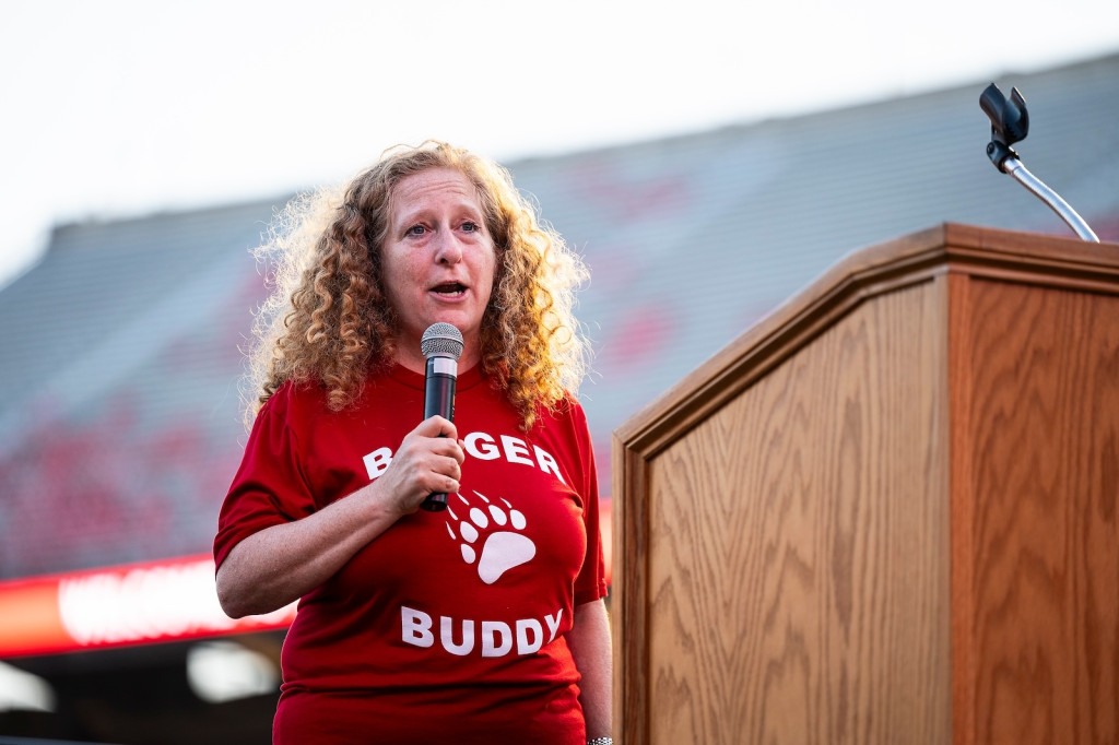 A woman with a red Badger Buddy T shirt talks at a podium.