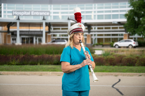 Photo of a woman wearing blue hospital scrubs and a marching band hat as she plays in front of a hospital entrance.