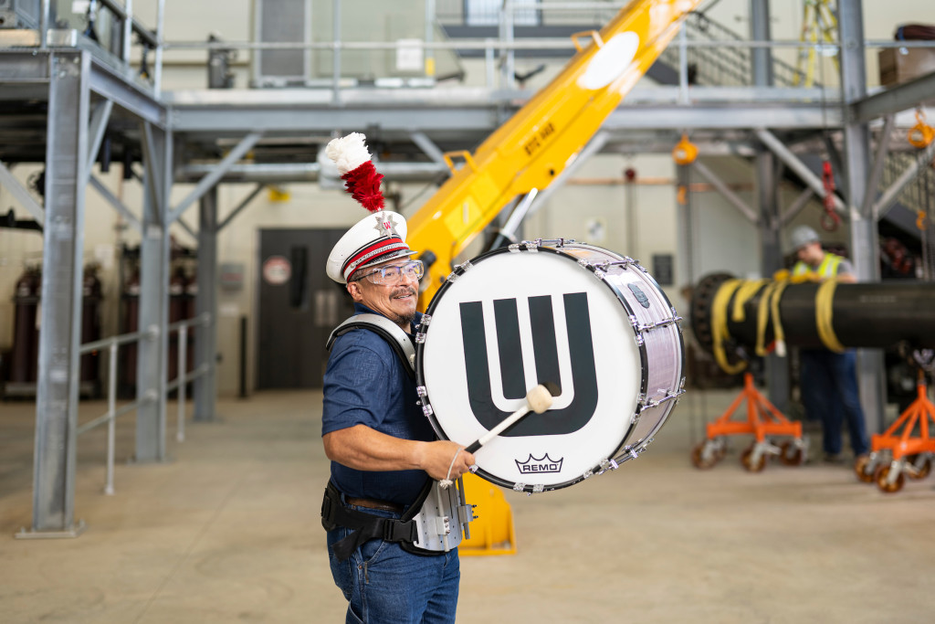 A man in a blue shirt, denim jeans and a marching band hat beats the bass drum strapped to his chest as he stands in an industrial facility.