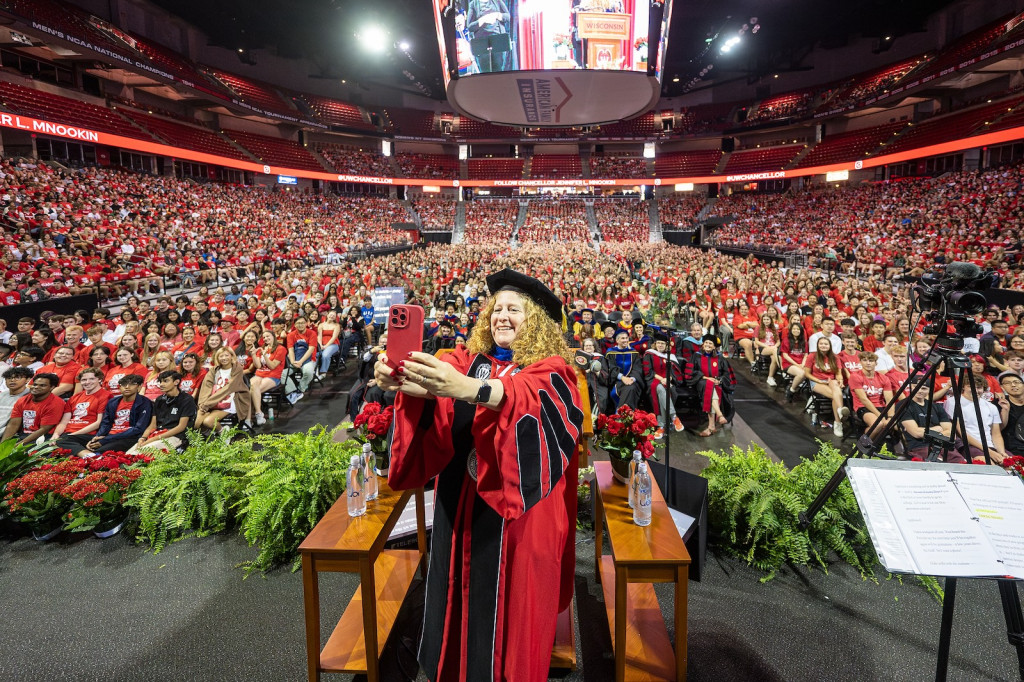 A woman at a podium in front of an arena full of people turns and takes a selfie of all of them.