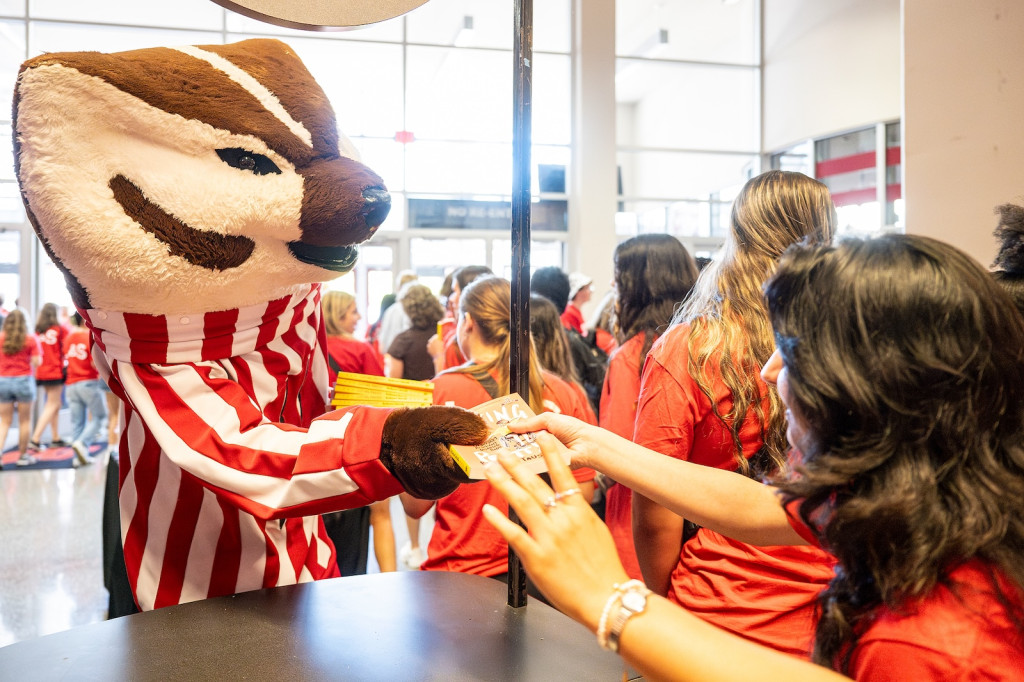 A person in a Bucky Badger costume hands out books to students.