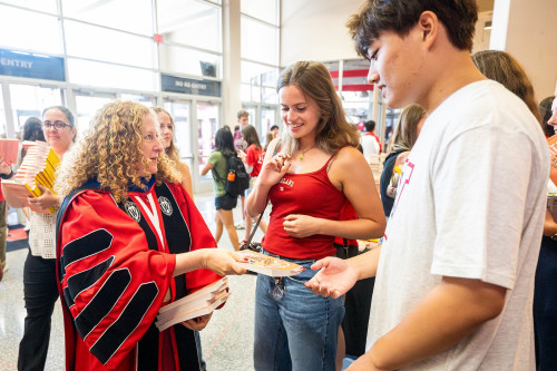 A woman in academic robes hands out books to students.