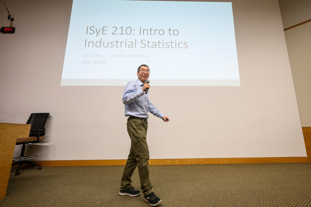 A professor stands on a stage in front of a projector screen.