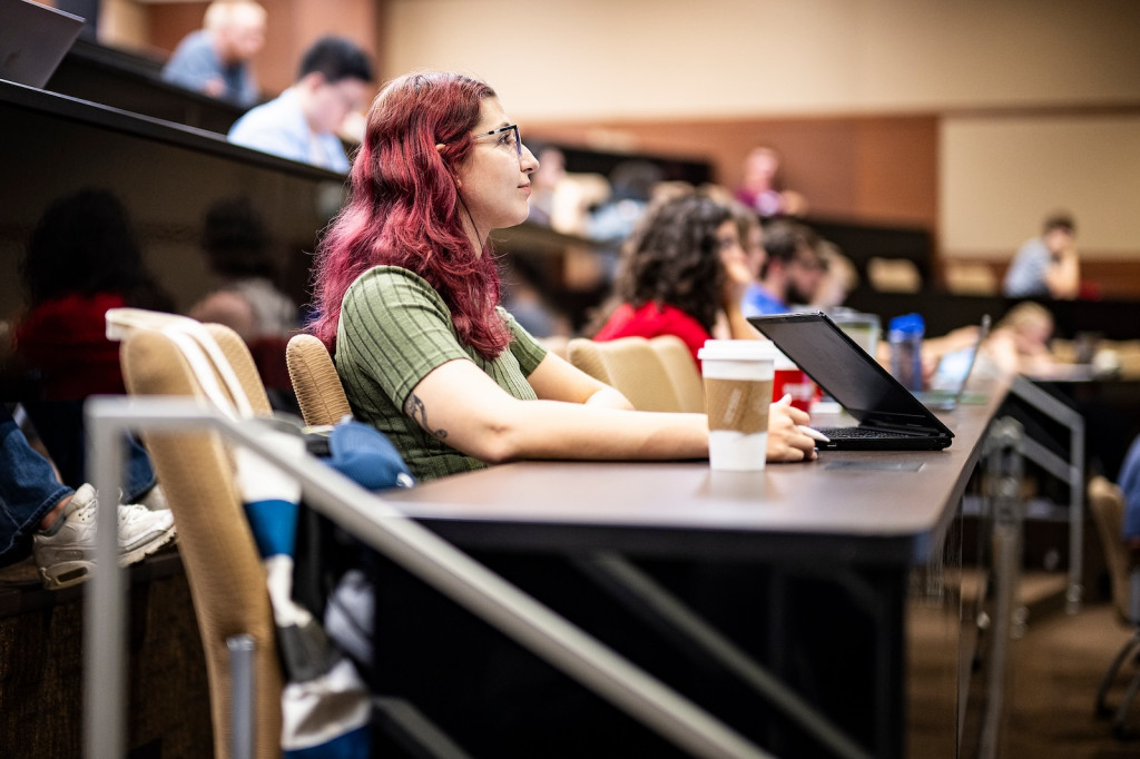 A woman sits at a desk in a lecture hall and listens.